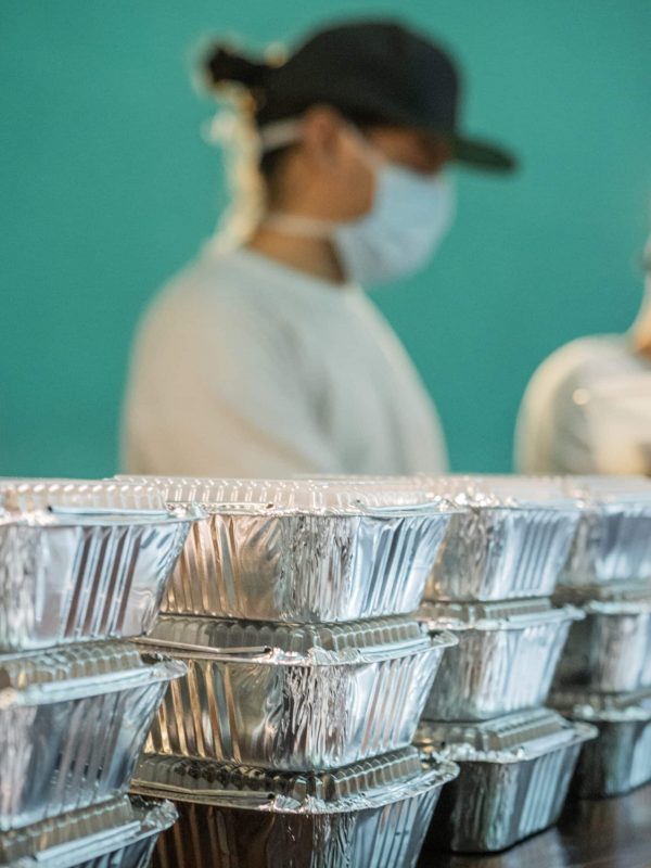Close-up of aluminum trays with food to deliver. Man with mouth cap in the background.